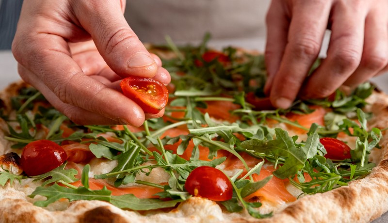 close-up-man-putting-tomatoes-baked-pizza-dough-with-smoked-salmon-slices (1)